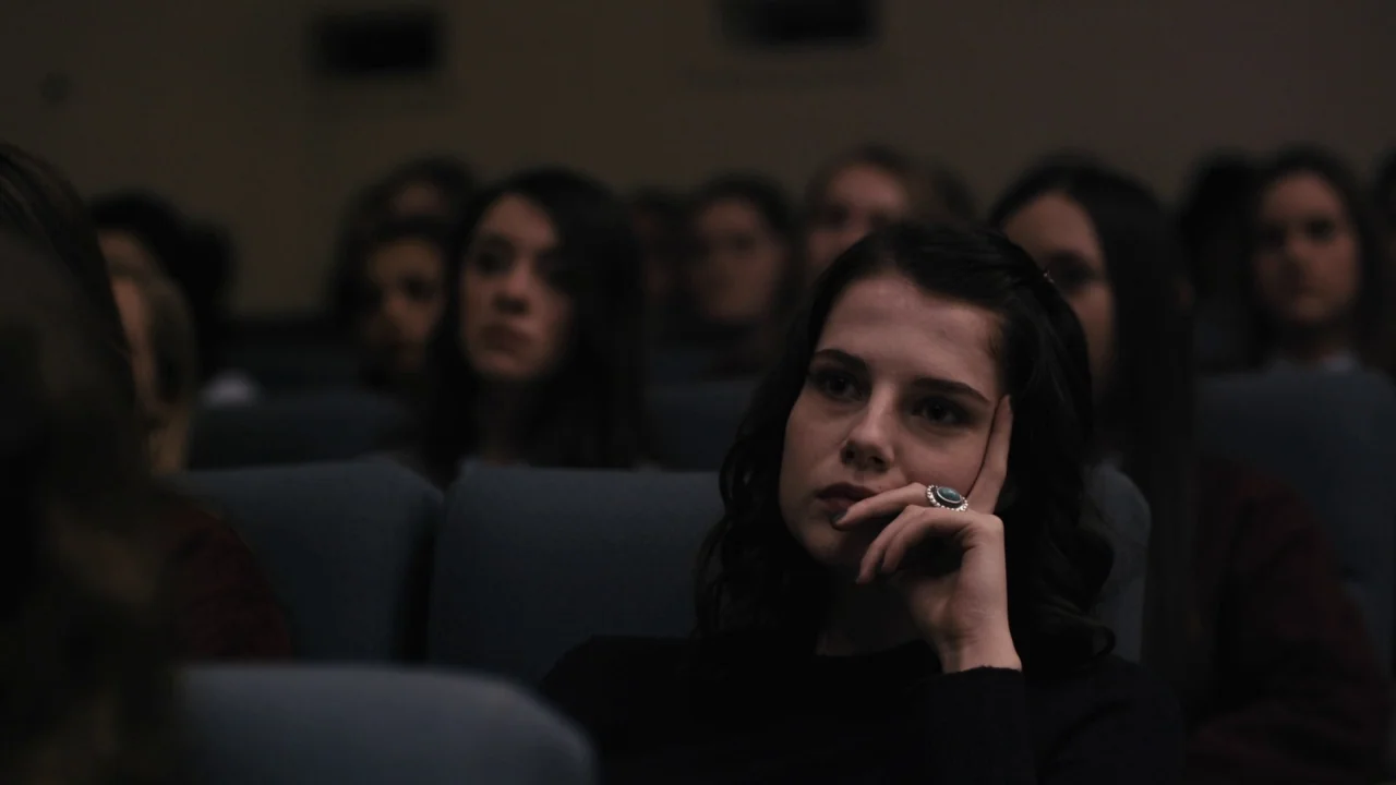 The Blackcoat’s Daughter (2015) movie still: medium — A dark auditorium with rows of blue padded seats occupied by an audience. A young woman in the…