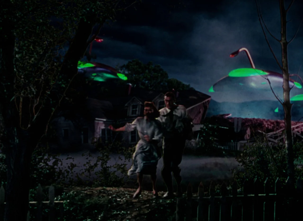 The War of the Worlds (1953) movie still: fullbody — A suburban house at night with dark, looming trees and a wooden picket fence. Two people run…