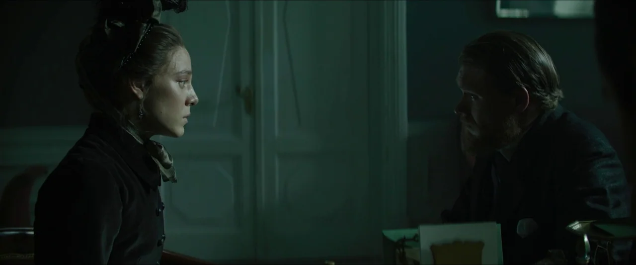 Tchaikovsky’s Wife (2022) movie still: medium — A dimly lit room with muted green tones and a panelled wooden door behind. A man and a woman face…