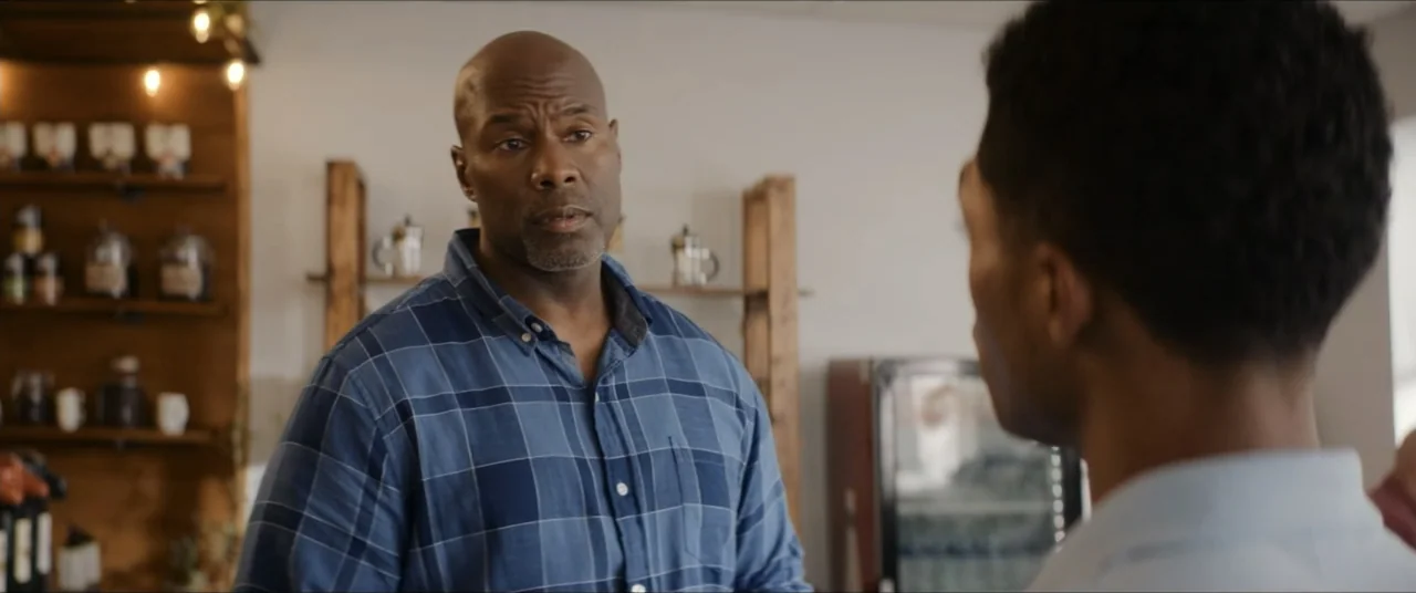The Forge (2024) movie still: medium — A well-lit interior of a cafe features wooden shelving units against a plain wall. A man in a blue plaid…