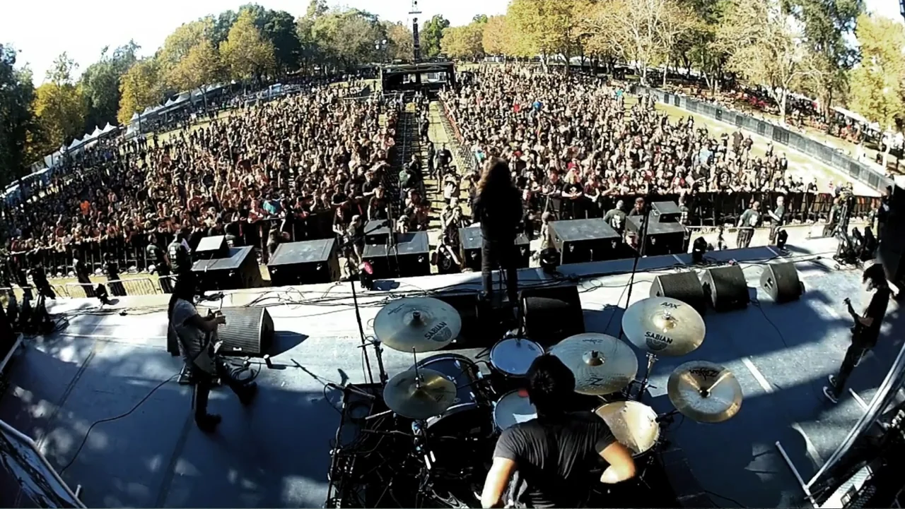 Control (2022) movie still: wide — An outdoor festival venue shows a band performing on stage before a massive crowd. The performers face a large…