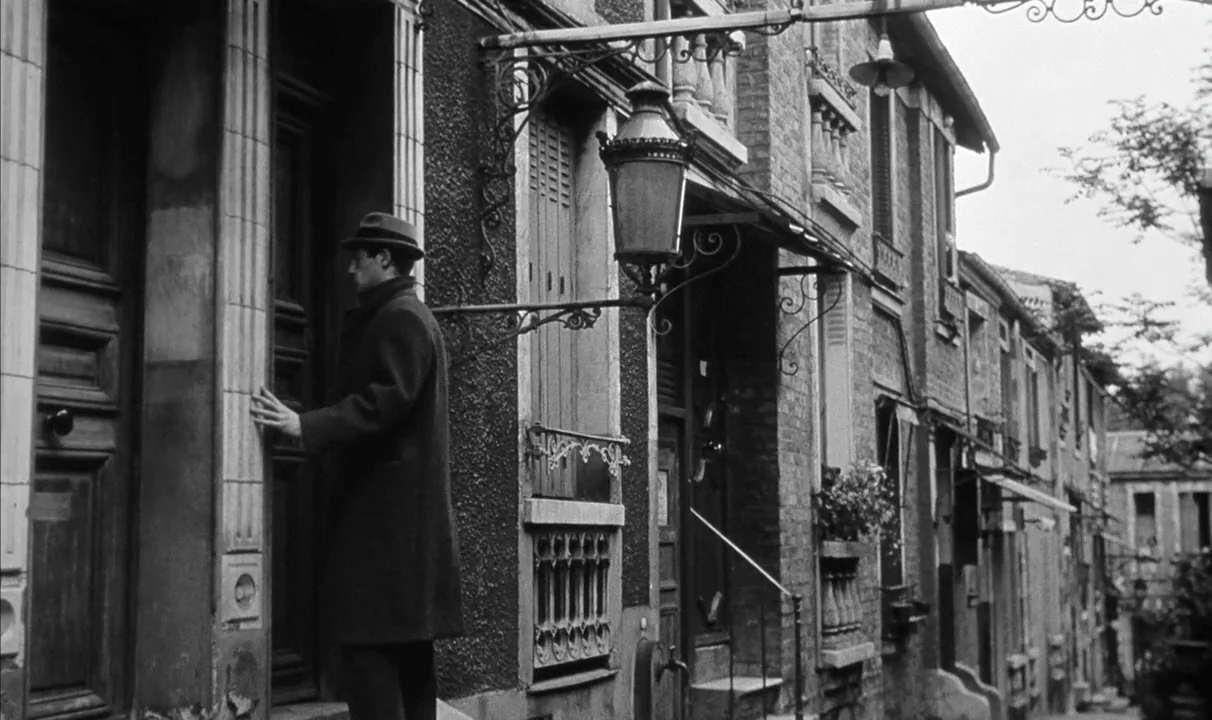 Le Doulos (1962) movie still: medium — A narrow residential urban street features brick masonry buildings with ornate iron lamp fixtures. A man…