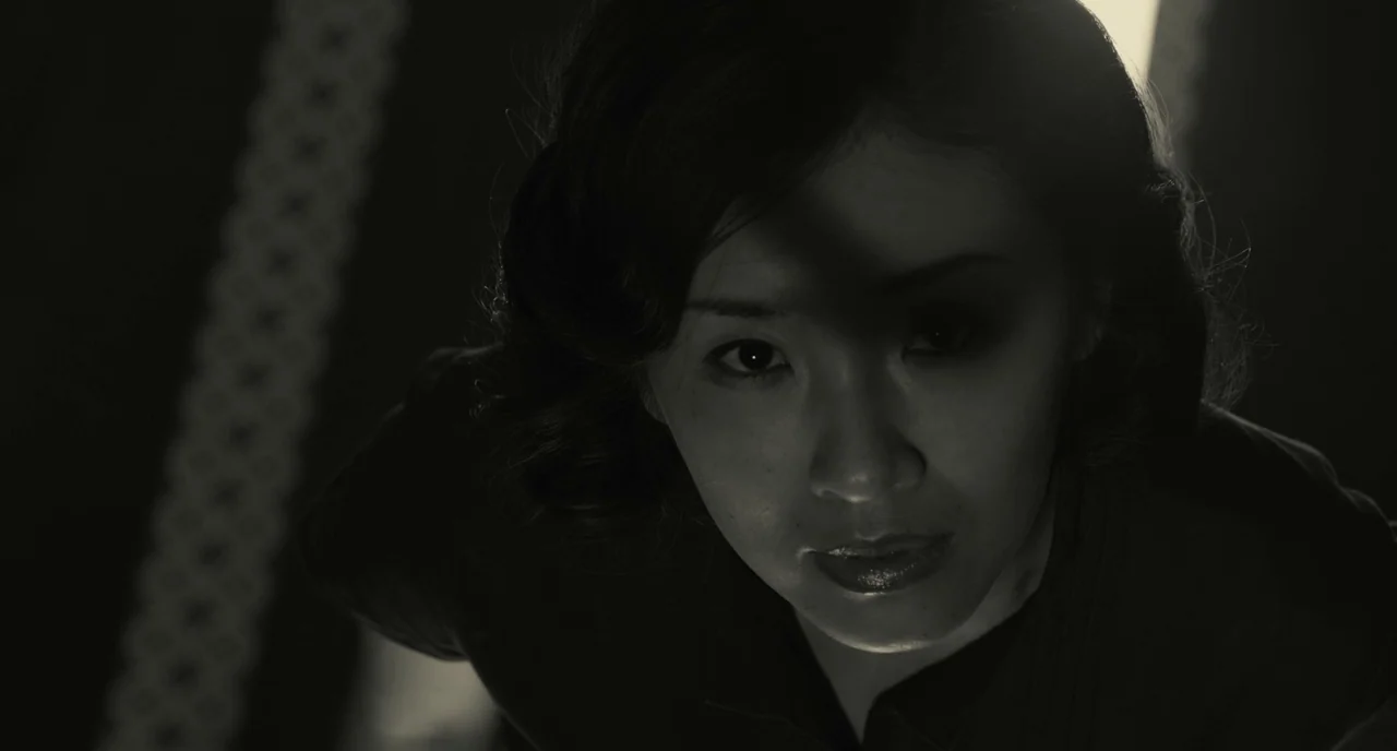 The Whispering Star (2015) movie still: closeup — A dimly lit indoor space features a dark room with a single light source. A woman gazes directly…