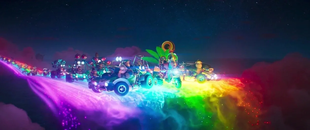 The Super Mario Bros. Movie (2023) movie still: wide — A group of ape-like characters drive small karts across a vibrant glowing rainbow track. The…