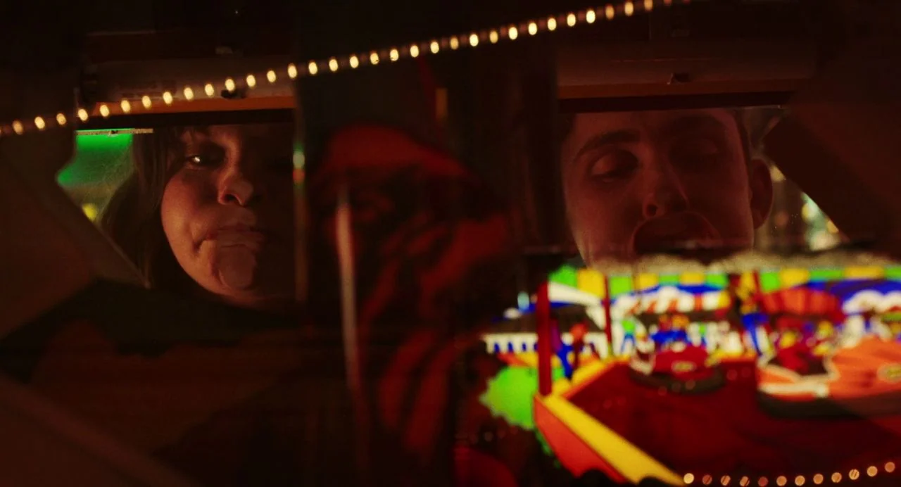 Dating Amber movie still: medium — Two people gaze downward into the glowing, brightly colored display of an arcade cabinet. The environment is…