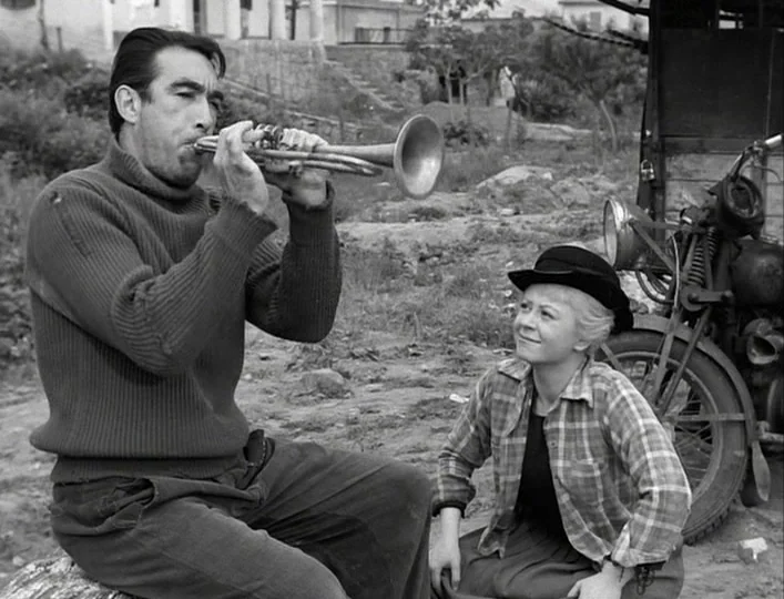 La Strada (1954) movie still: medium — An open dirt field with an old motorcycle and stone wall in the background. A man plays a trumpet while a…