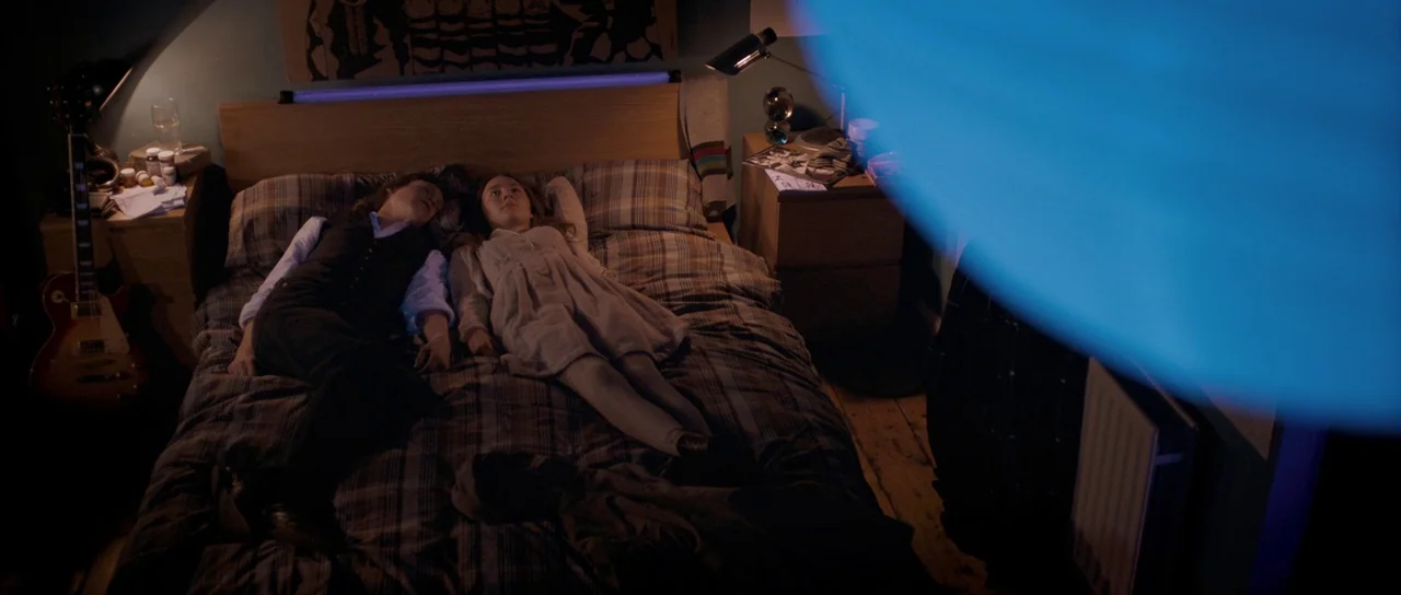 Byzantium (2012) movie still: wide — A cluttered bedroom is illuminated by a glowing blue tube light above the headboard. Two teenagers lie flat on…