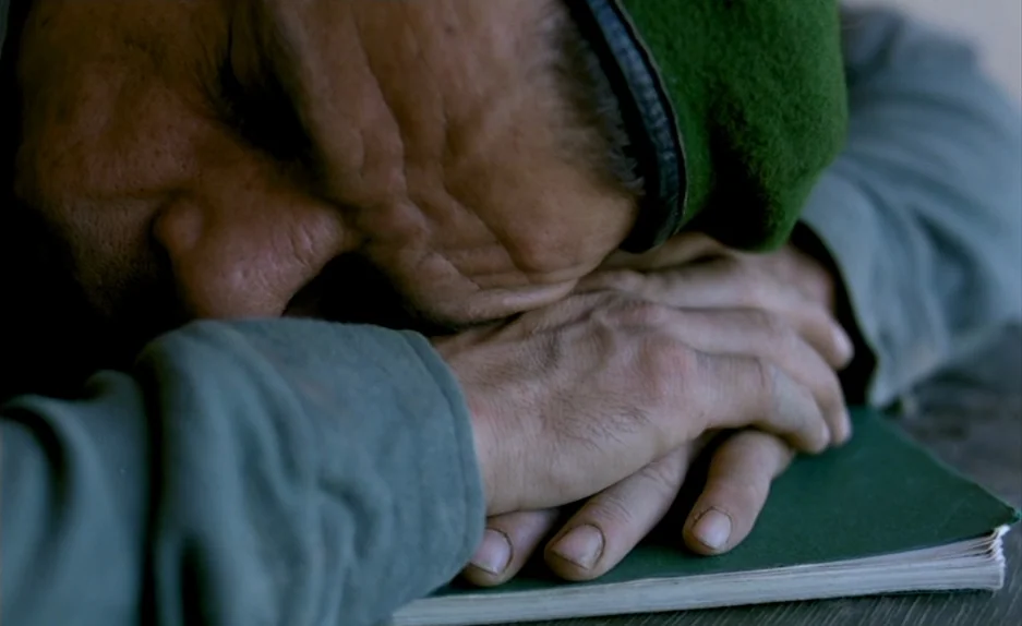 Beau Travail (1999) movie still: closeup — A man sleeps with his face pressed against his hands over a green book. The setting is minimal with muted…