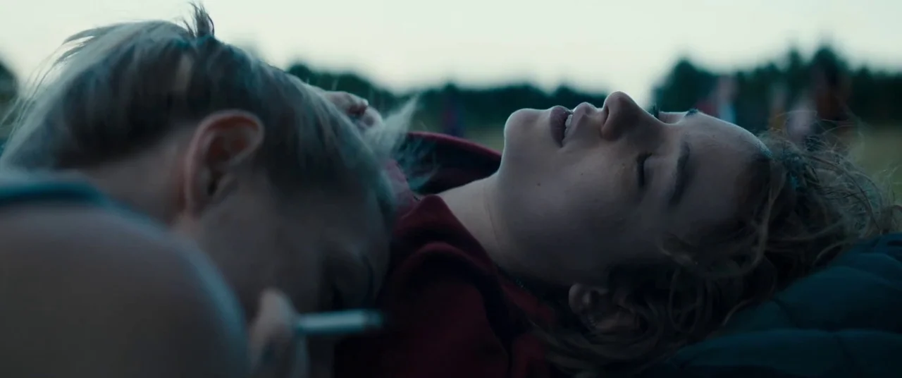 Silver Haze (2023) movie still: closeup — Two young women lie on their backs in a grassy field under a dim sky. Their heads are positioned close…