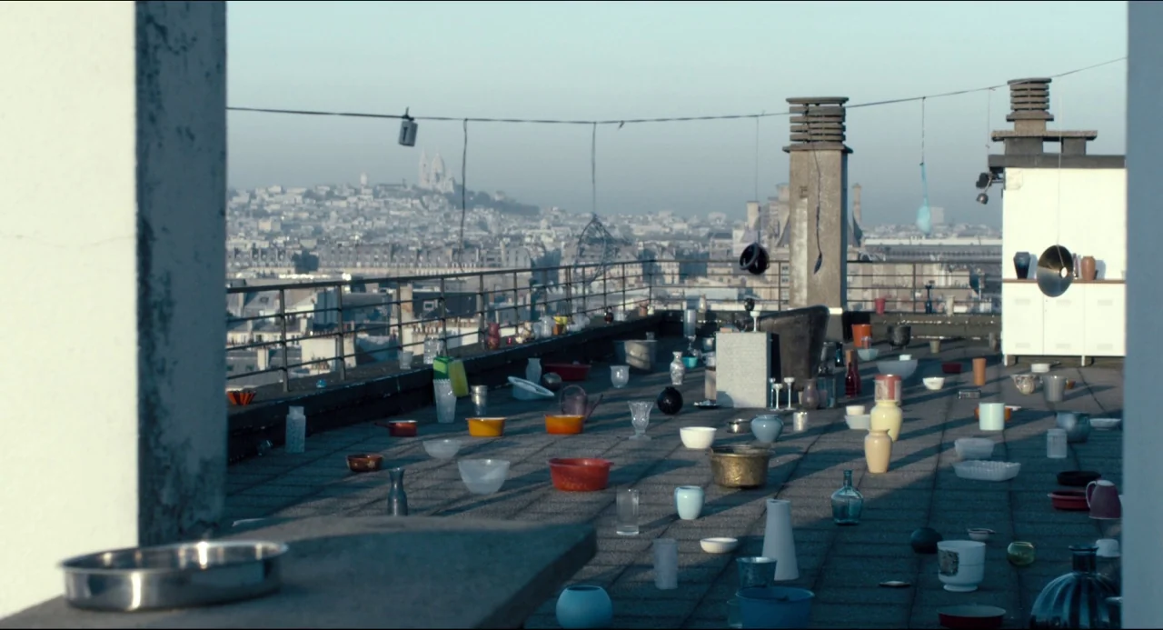 The Night Eats the World (2018) movie still: wide — A rooftop terrace paved with concrete tiles overlooks a distant cityscape under pale sky. Dozens…