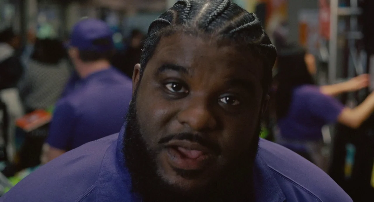 Trap (2024) movie still: closeup — A retail environment contains rows of merchandise partially visible in the blurred background. A man in a purple…