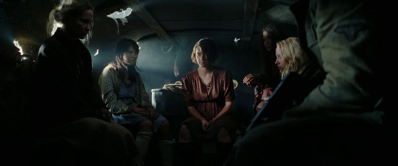 Sisu (2022) movie still: wide — The interior of a dark, cramped truck canopy with sunlight filtering through roof tears. Five women sit on wooden…