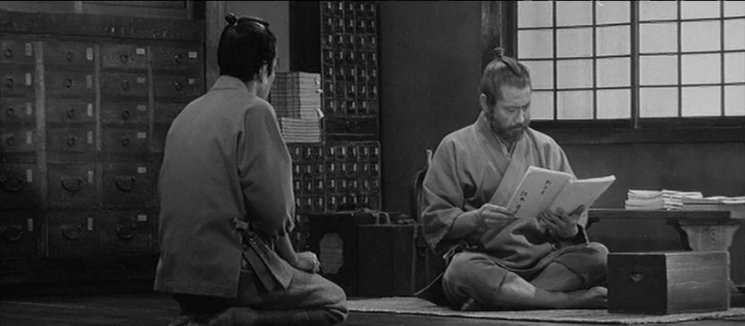 Red Beard (1965) movie still: medium — A Japanese apothecary interior features rows of wooden medicinal cabinets and a shoji window. One man sits…