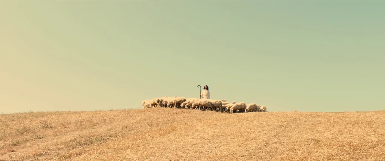 Benedetta (2021) movie still: wide — A sun-drenched, dry grassy landscape under a clear pale sky. A man in robes stands centrally among a large…
