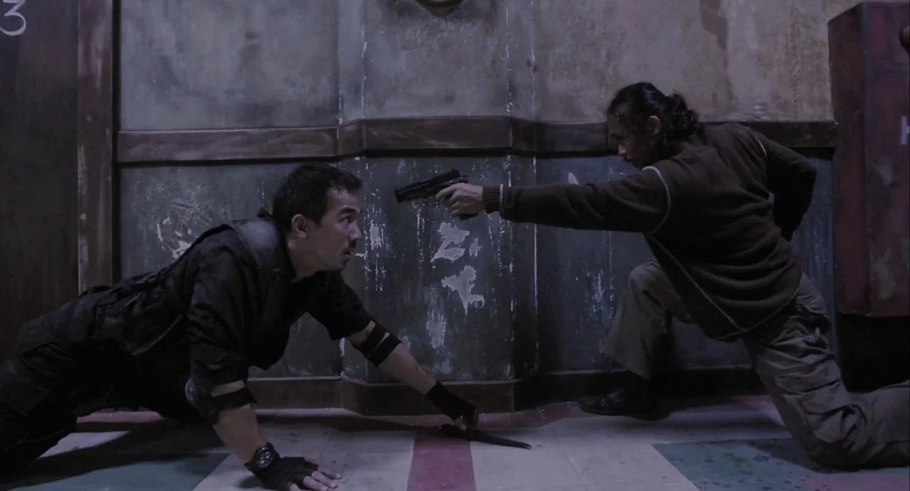 The Raid (2012) movie still: fullbody — A dimly lit, concrete hallway with faded wall markings and institutional paint. Two men are engaged in a…