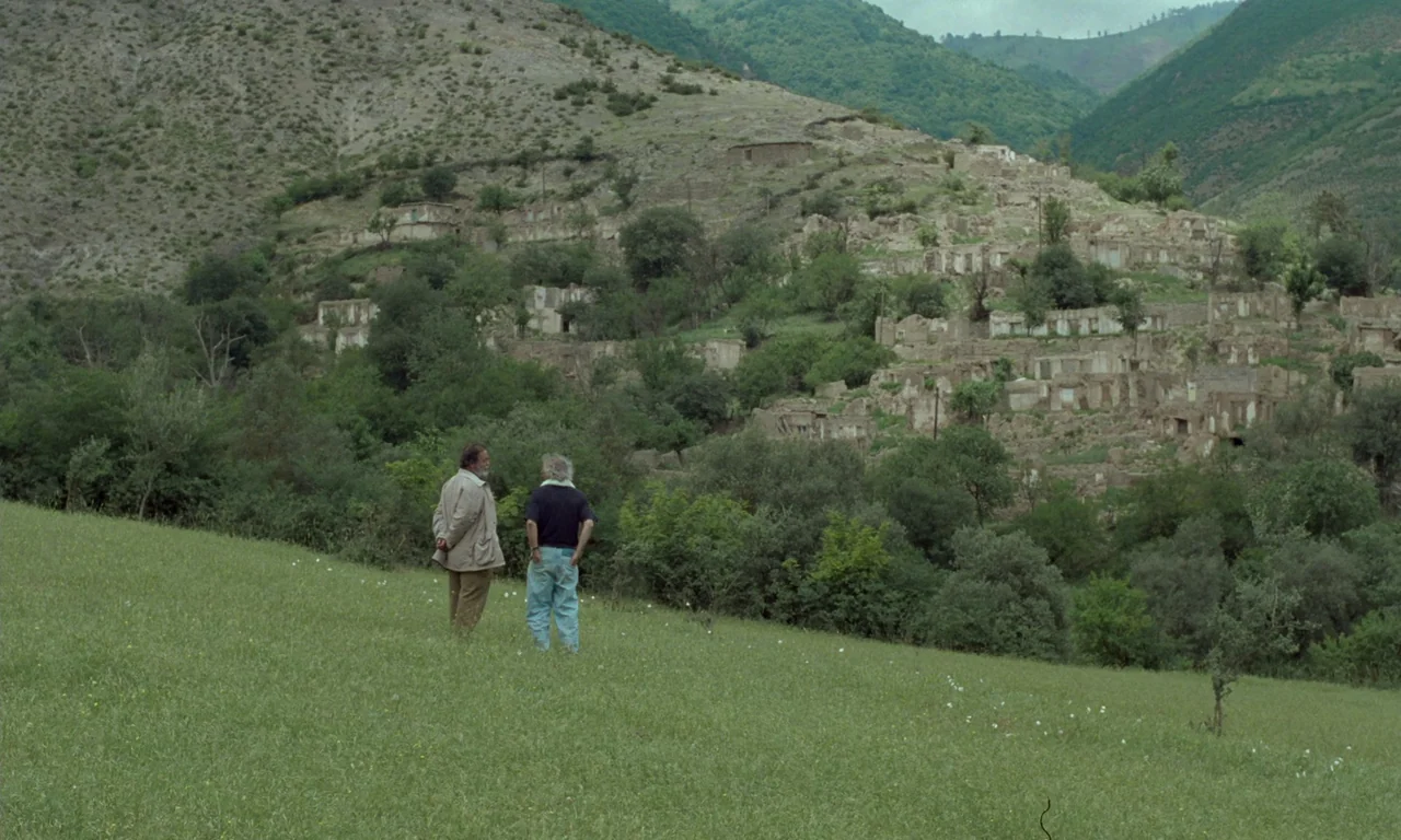 Through The Olive Trees (1994) movie still: wide — A green hillside overlooks a cluster of ruined stone buildings in a valley. Two men stand in the…