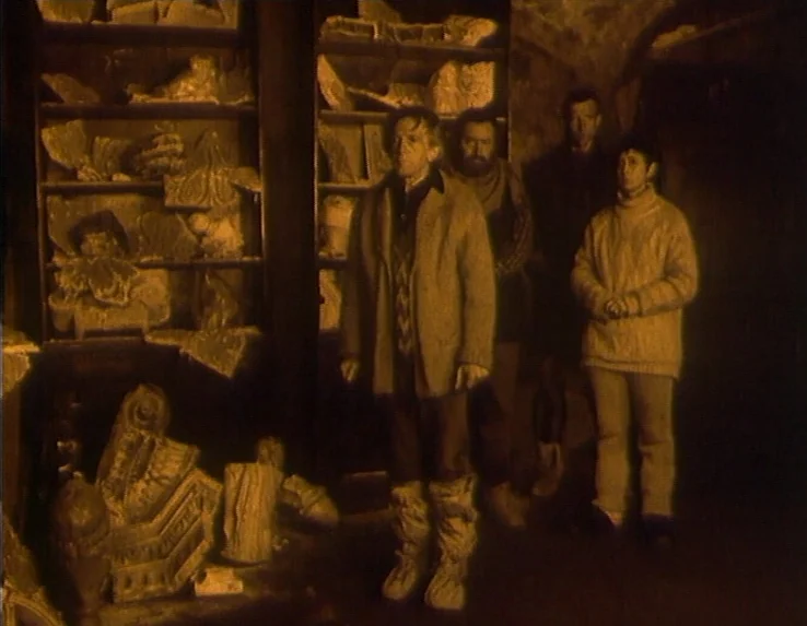 Dead Man’s Letters (1986) movie still: fullbody — A dimly lit storage area filled with shelves containing broken stone architectural fragments. Four…