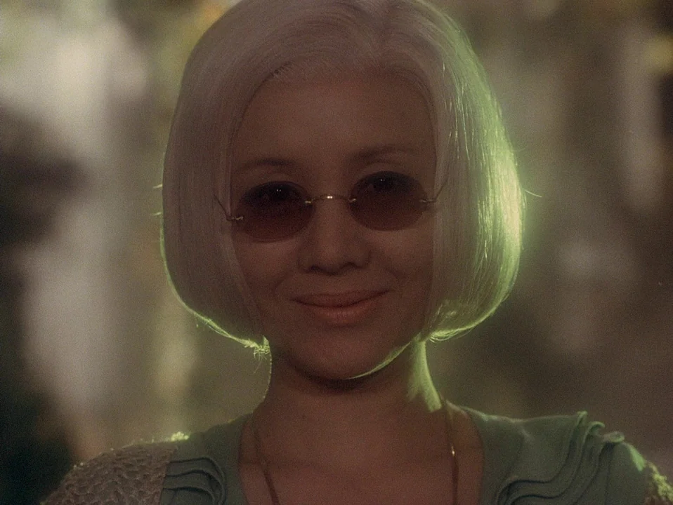 House (Hausu) (1977) movie still: closeup — A woman stands in a dimly lit interior space with soft green illumination. She wears sunglasses and a…