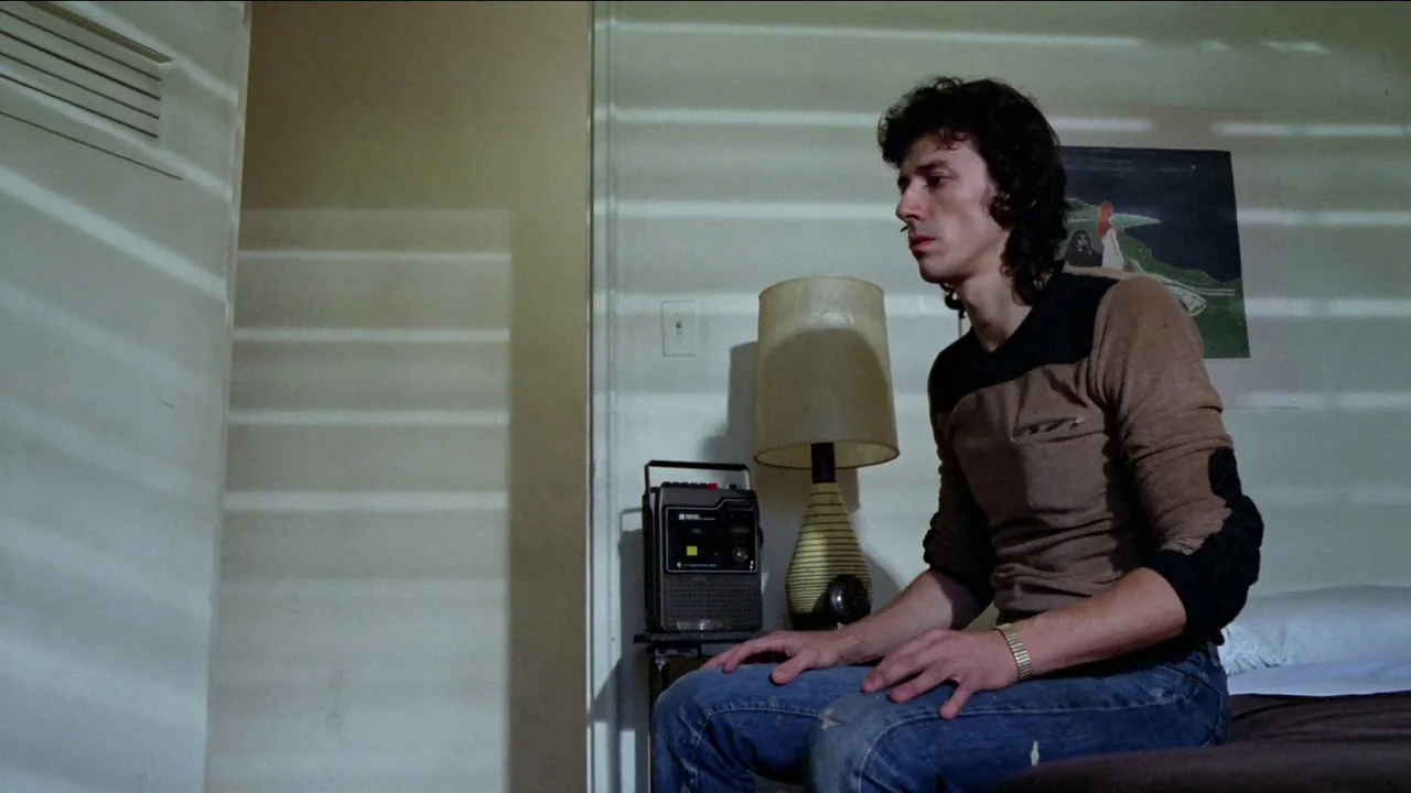 Cruising (1980) movie still: medium — A spare room with white walls, a bedside lamp, and a portable radio. A man sits motionless on the bed while…