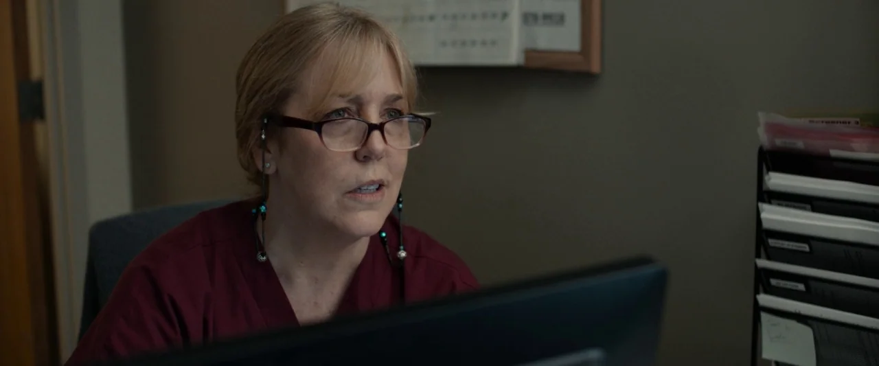 Little Woods (2018) movie still: medium — A medical office environment with a document rack and blurred papers visible. A woman in burgundy scrubs…