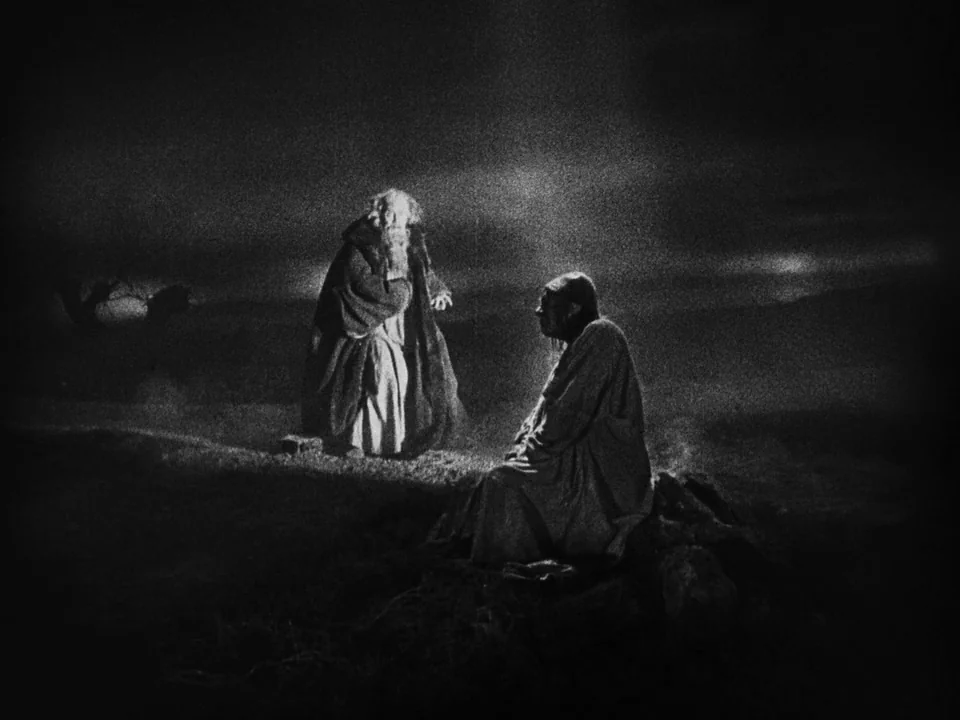 Faust (1926) movie still: wide — A dark, open field serves as the backdrop for two figures at night. A bearded man in robes stands while another man…