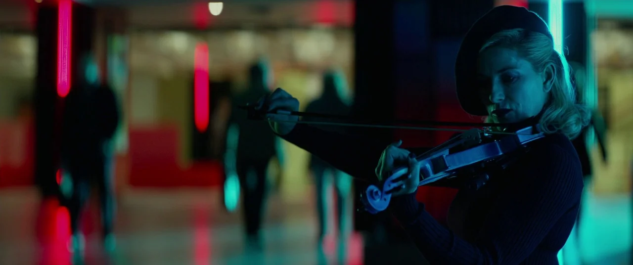 John Wick: Chapter 2 (2017) movie still: medium — A dark corridor features blurred figures walking in the background under glowing neon lights. A…