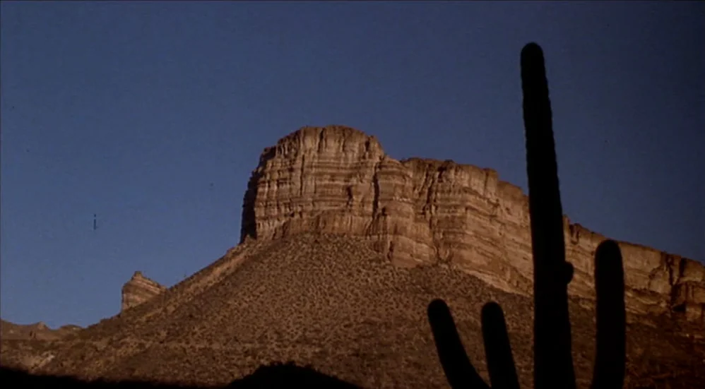U-Turn (1997) movie still: establishing — A tall mesa with layered sandstone cliffs rises against a clear blue sky. A large saguaro cactus is…