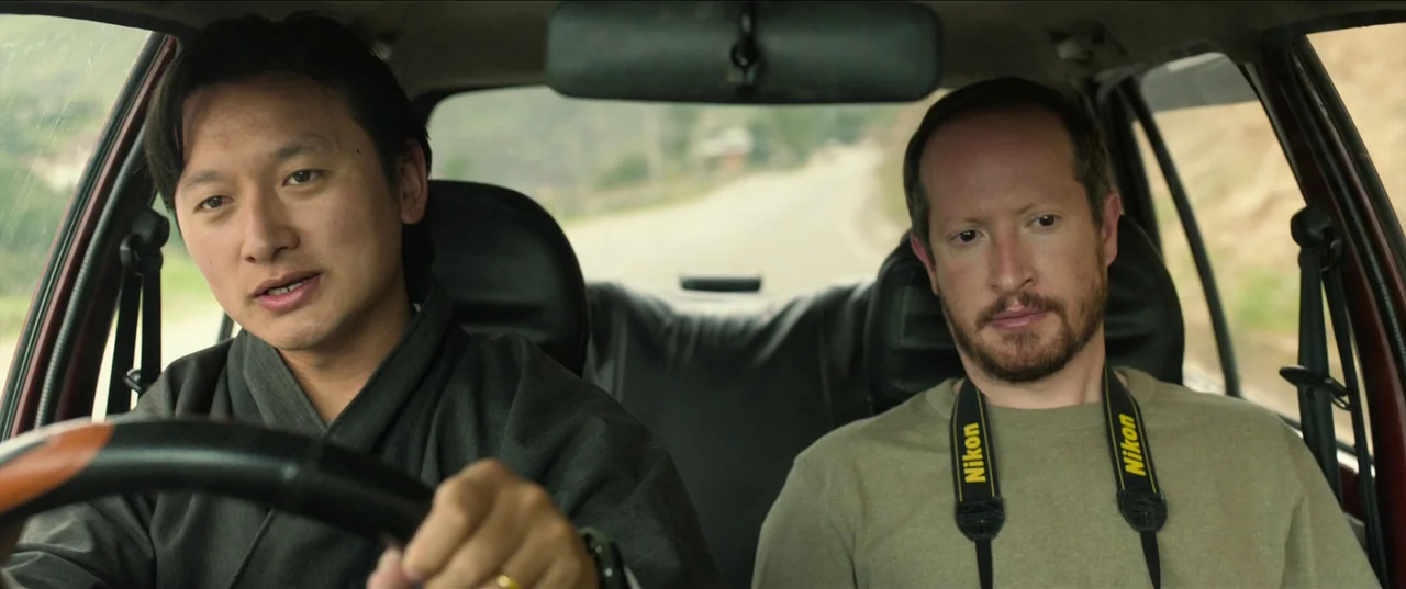 The Monk and The Gun (2023) movie still: medium — Two men sit in the front seats of a car moving along a rural road. The driver holds the steering…