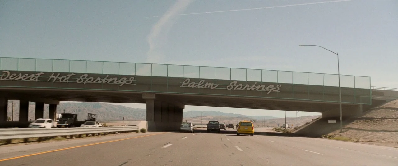 Destroyer (2018) movie still: wide — An arid desert highway passes underneath a large concrete bridge with prominent metal signage. Several…