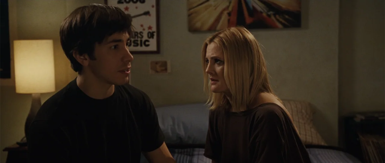 Going the Distance (2010) movie still: medium — A bedroom contains a bed with a blue blanket and a small bedside lamp. A man and a woman are seated…
