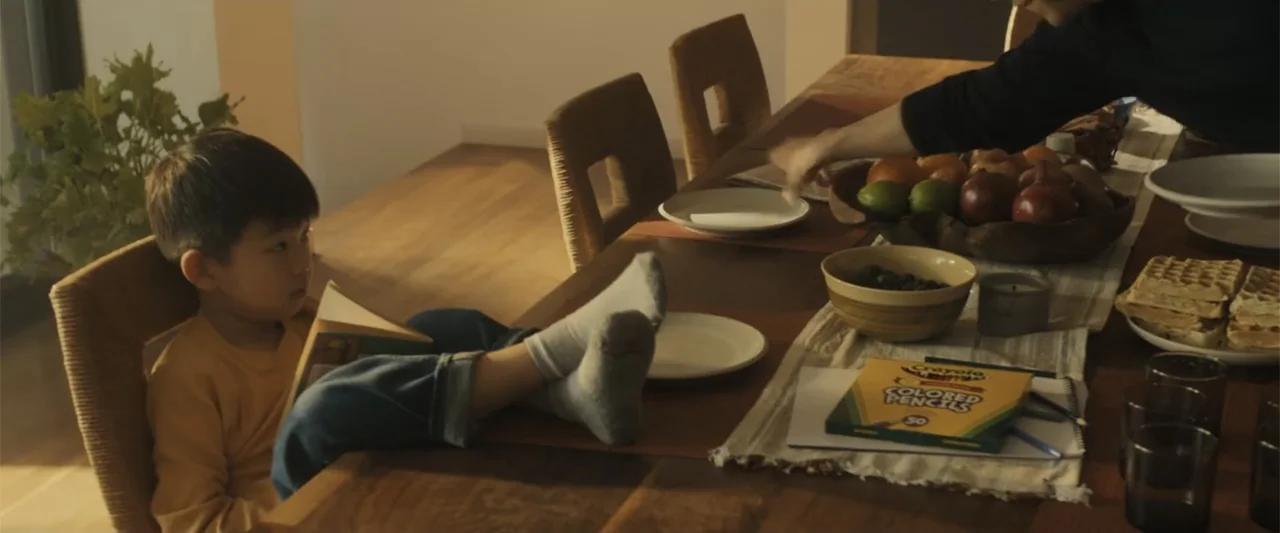 Afraid (2024) movie still: medium — A wooden dining table features a bowl of fruit, a box of colored pencils, and plates. A young boy sits in a…