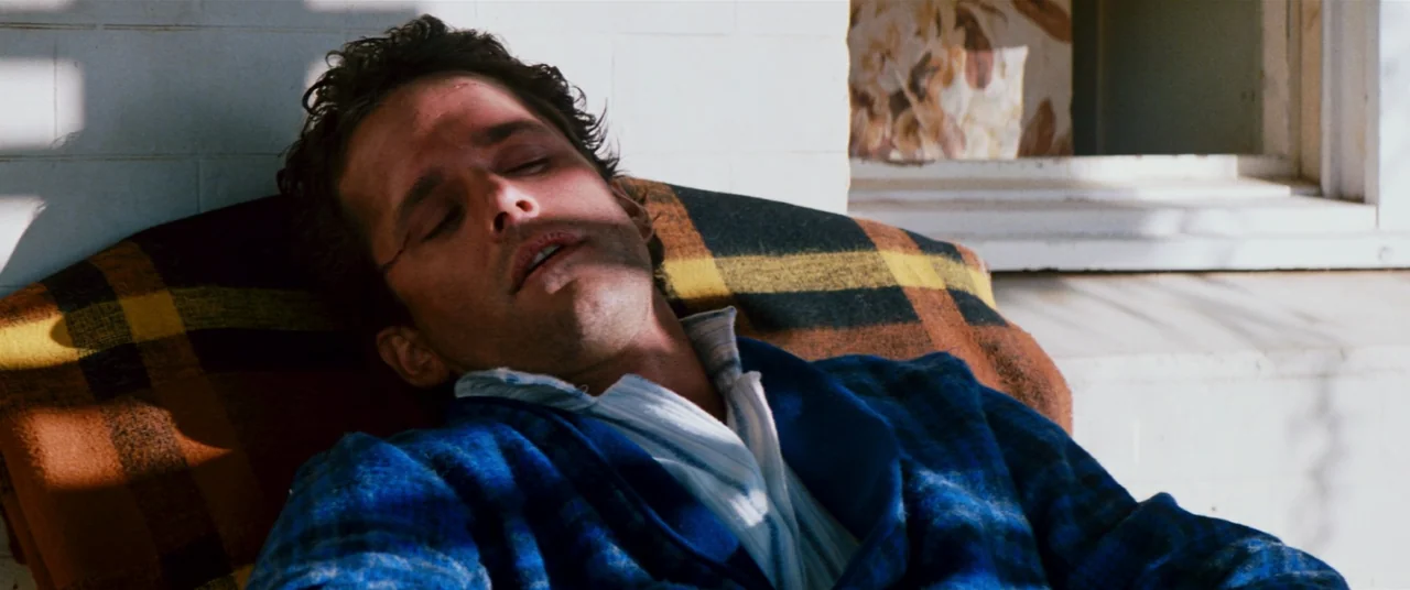 Razorback (1984) movie still: closeup — A person rests on a plaid cushion against a white painted masonry wall. The sunlight creates distinct…