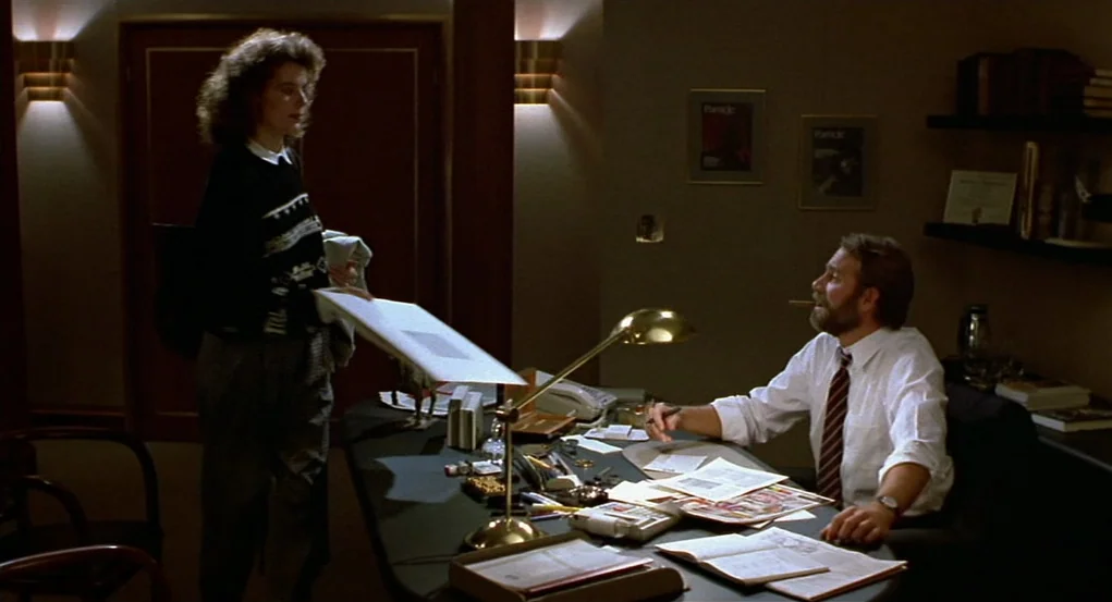 The Fly (1986) movie still: medium — An office at night with dark wood paneling and warm wall sconce lighting. A man sits at a desk cluttered with…