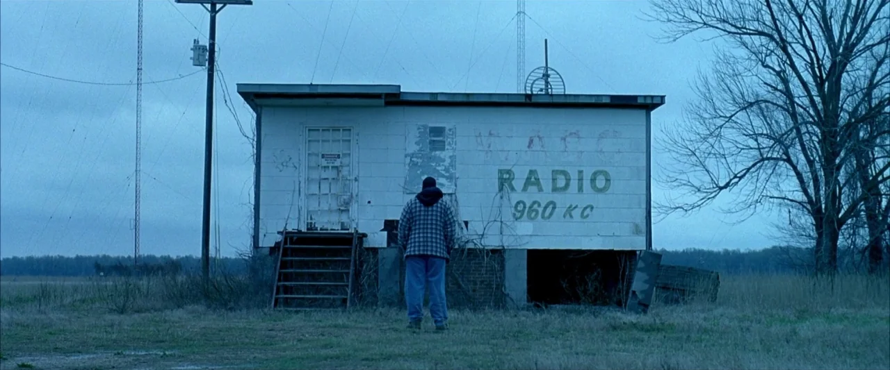 Ballast (2008) movie still: wide — An isolated, weathered concrete radio station stands in a barren, grassy field at twilight. A lone figure wearing…