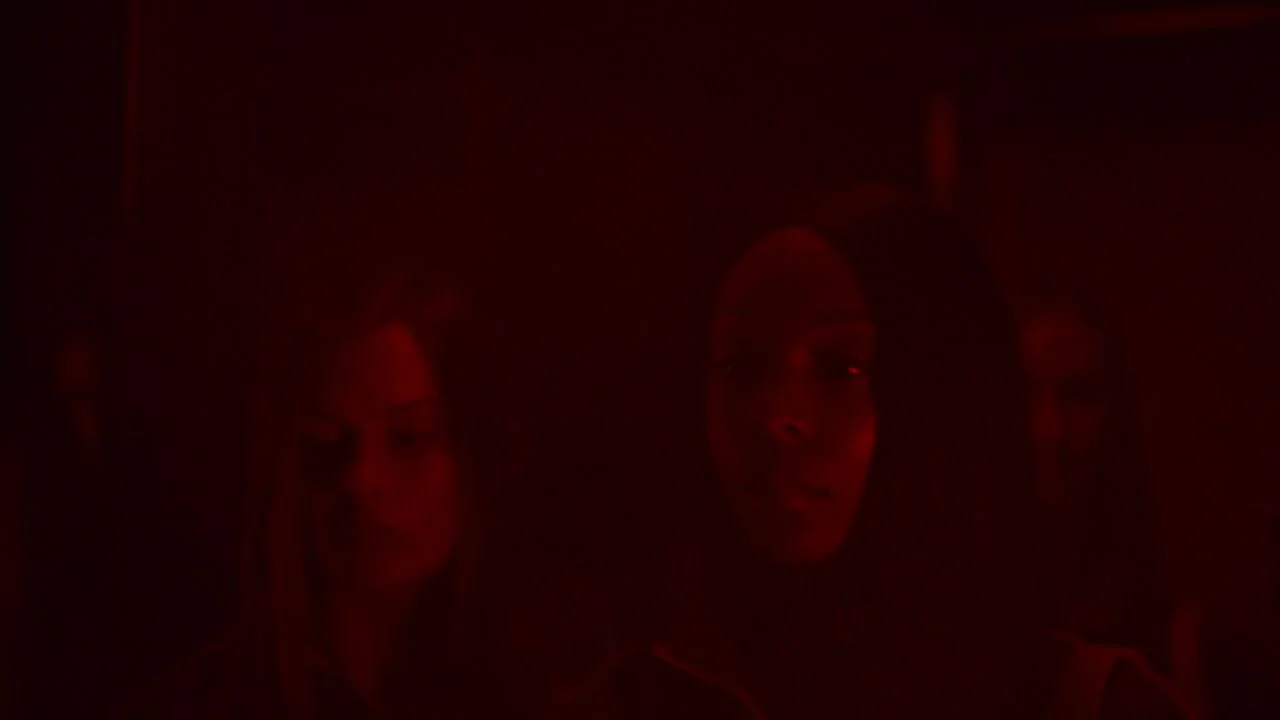 Possessor (2020) movie still: medium — A dimly lit room is bathed in thick, saturated red light. Three individuals stand close together in the dark…