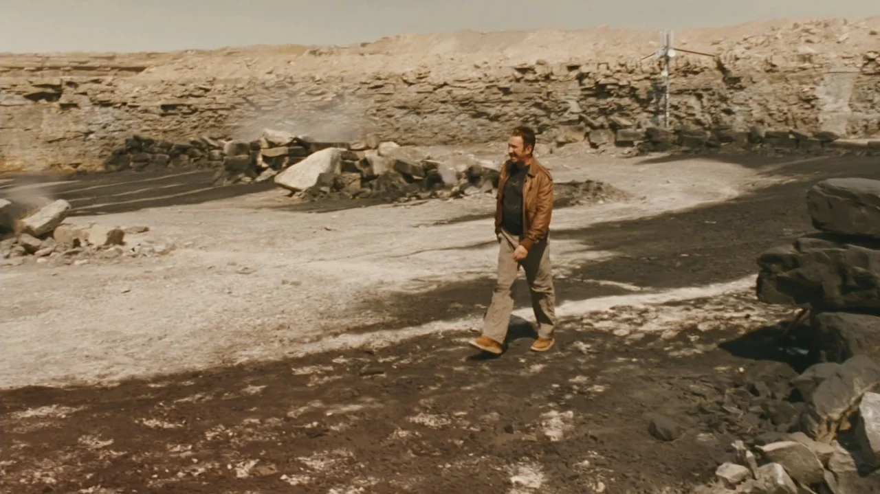 Zoom (2006) movie still: fullbody — An arid stone quarry features layered rock walls and a dusty, dark-soiled ground surface. A man walks across the…