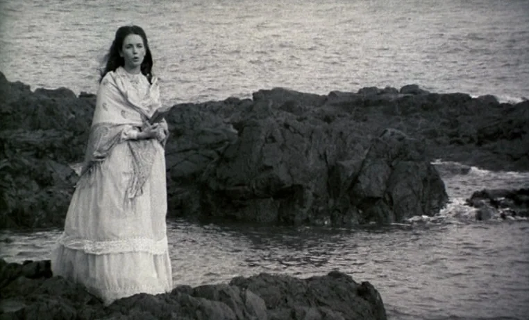 The Story of Adele H (1975) movie still: fullbody — A woman in a historical dress stands on jagged volcanic rocks near the ocean. She is positioned…