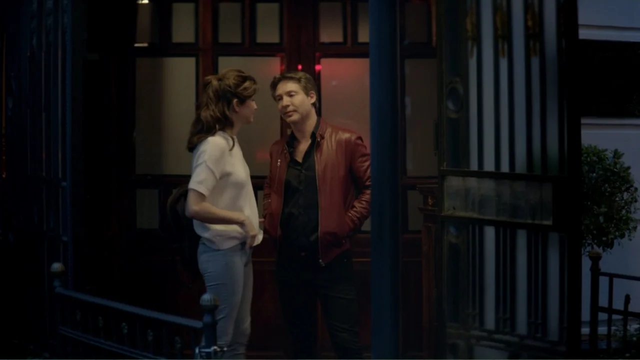 I Married a Dumbass (2016) movie still: medium — Two people stand outside a building with glass doors and a dark metal gate. A woman in a white…