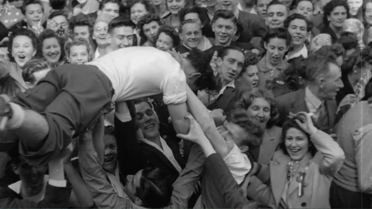 The Witch of Kings Cross (2020) movie still: medium — A dense crowd of people in 1940s clothing celebrate outdoors in an urban space. A person in a…
