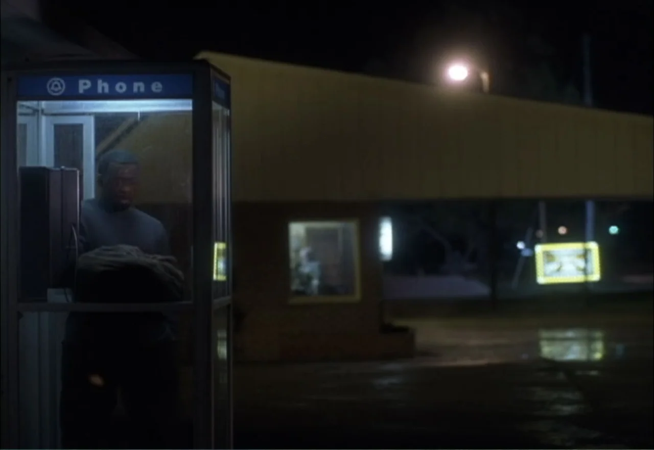 Ambushed (1998) movie still: medium — A man stands within an illuminated glass telephone enclosure during the night. The surrounding dark asphalt…