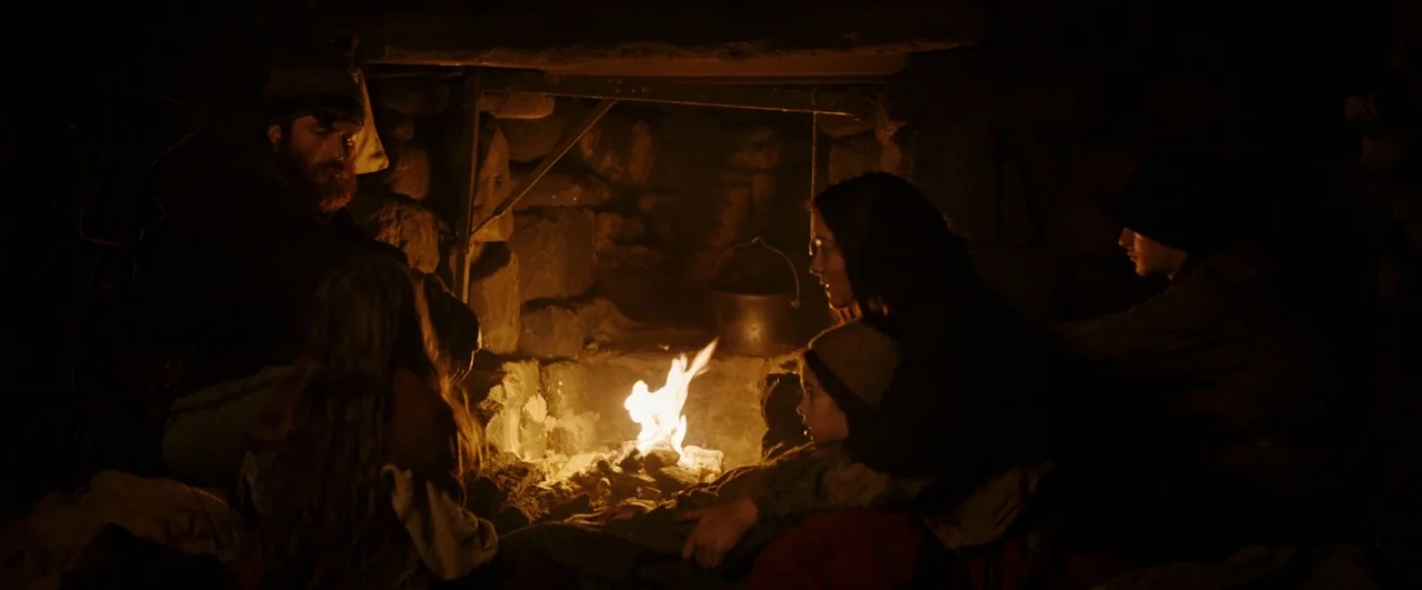 Black ’47 (2018) movie still: wide — A dark, rustic interior space features a rough-hewn stone fireplace with a metal pot. A family of four huddles…