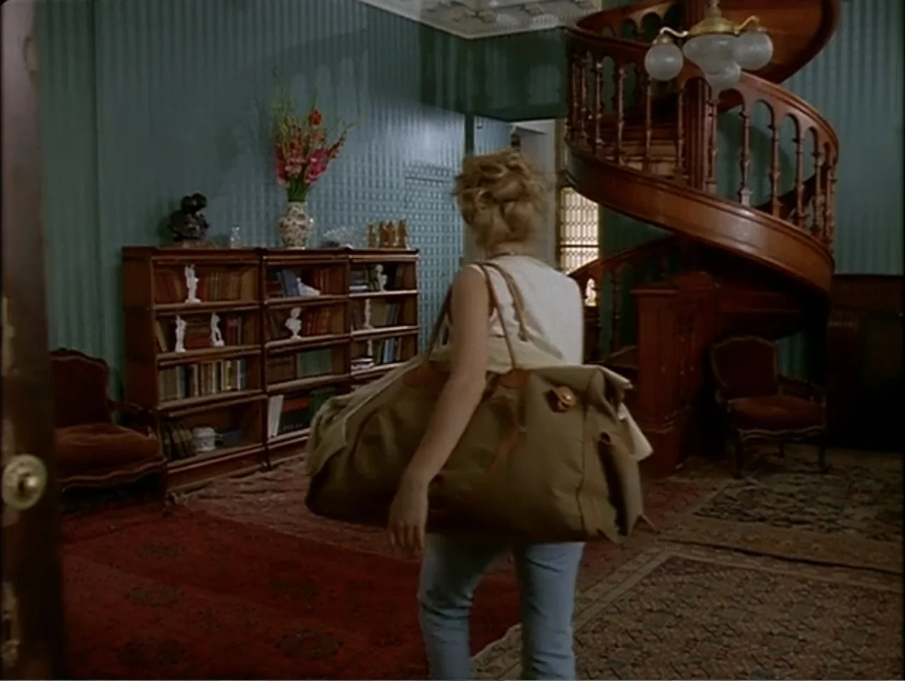 A Change of Place (1994) movie still: medium — A living room features green wallpaper, a spiral wooden staircase, and a wooden bookcase. A woman…