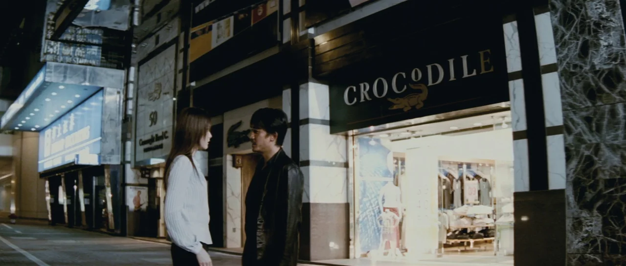 Infernal Affairs (2002) movie still: fullbody — A night view of a brightly lit storefront featuring a large Crocodille sign. A man and a woman stand…