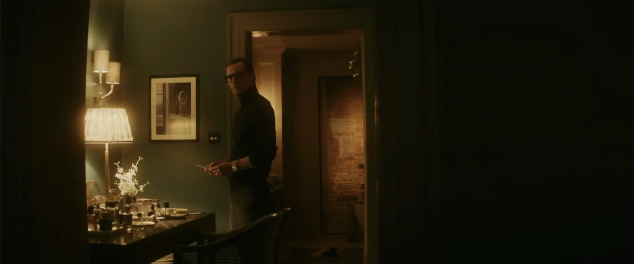Black Bag (2025) movie still: medium — A dark, dimly lit bedroom with teal walls features a vanity table and lamp. A man in a black turtleneck…