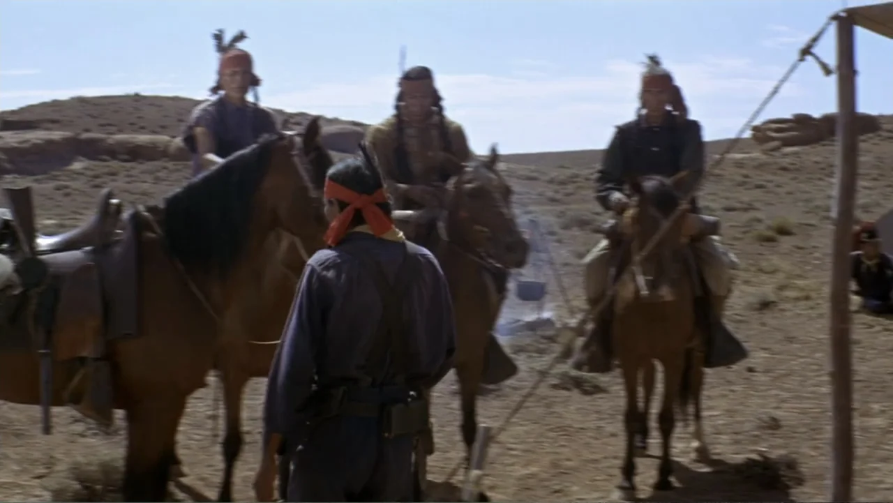 A Distant Trumpet (1964) movie still: wide — A dry, sun-baked desert encampment features sparse shrubs and uneven dirt ground. Four men mounted on…