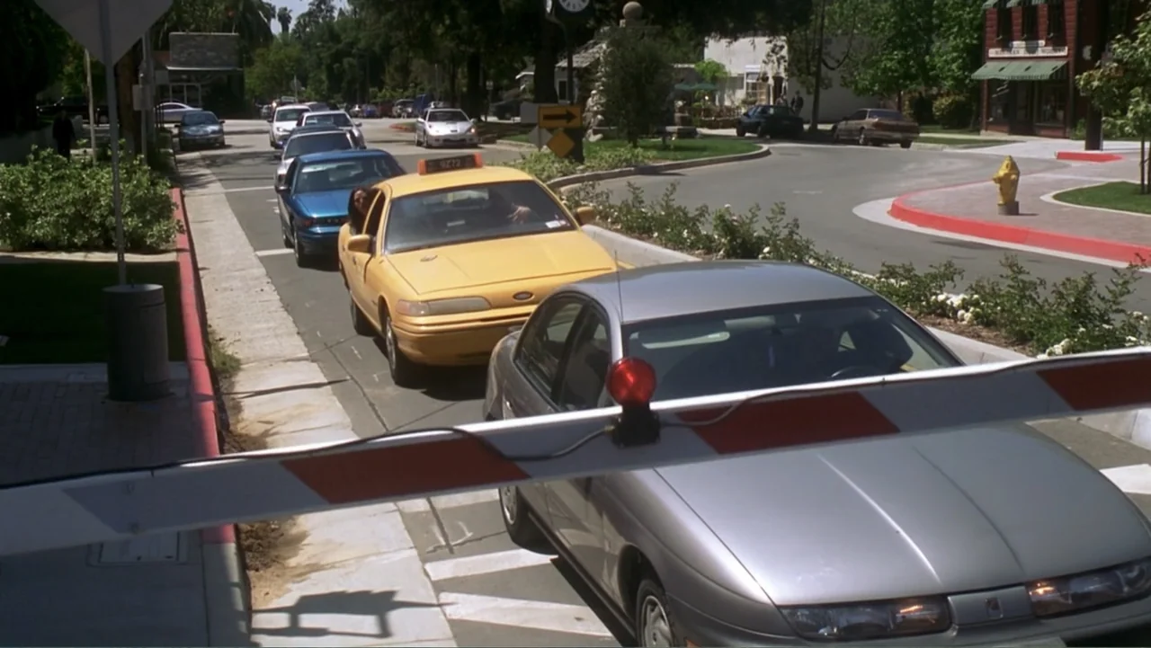 13 Going on 30 (2004) movie still: wide — A suburban entrance with a red and white barrier gate blocking a paved street. Several cars including a…