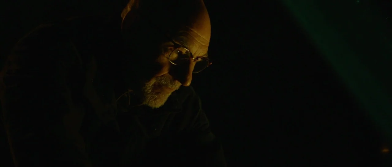 Green Room (2015) movie still: closeup — An elderly man is seen in a dark room with minimal ambient lighting. The subject looks downward while…