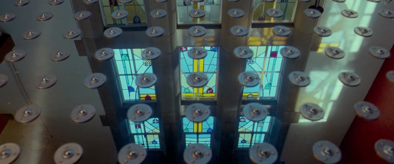 Catch Me Daddy (2014) movie still: establishing — A ceiling features a regular grid of metal rivets over reflected stained glass windows. These…