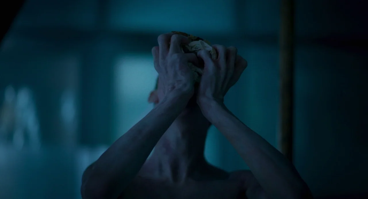 All You Need Is Death (2023) movie still: closeup — A dimly lit room with cool tones obscured in deep blue shadows. A shirtless individual holds a…