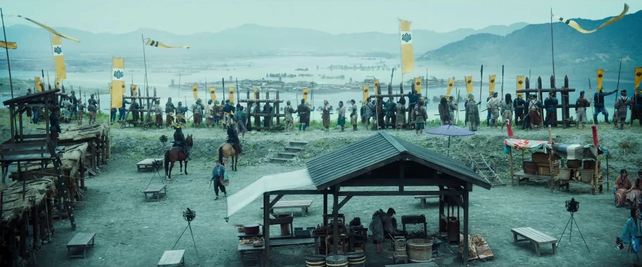 Kubi (2023) movie still: wide — A sprawling military camp with wooden structures and banners overlooks a misty lake landscape. Numerous soldiers in…